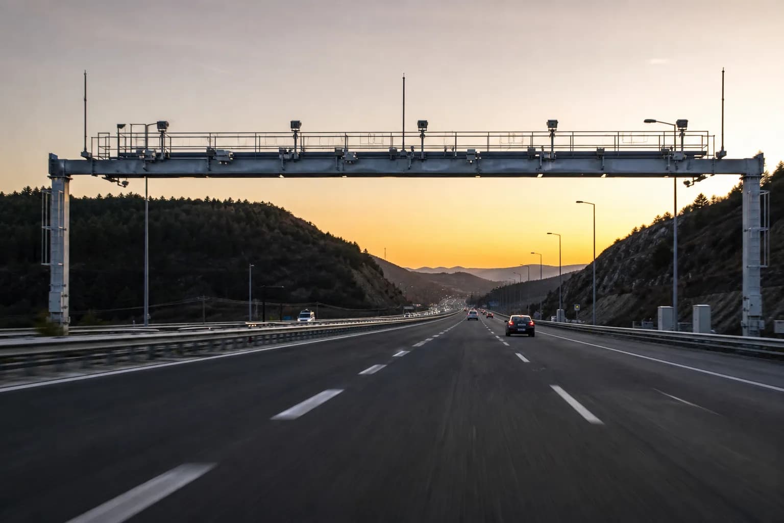 Gün batımında bariyersiz serbest geçiş portalının altından geçen otoyol; kameralı üst yapı şeritleri boydan boya kaplıyor.