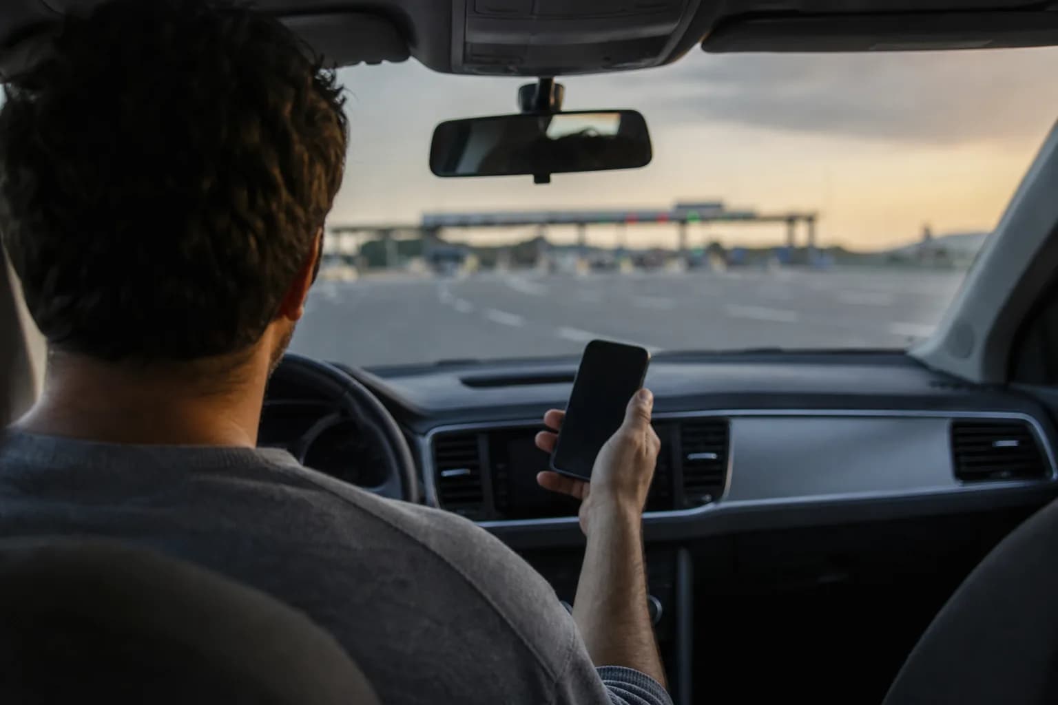Fahrer mit Smartphone im geparkten Auto an einer Raststätte, Mautstraße im Hintergrund.