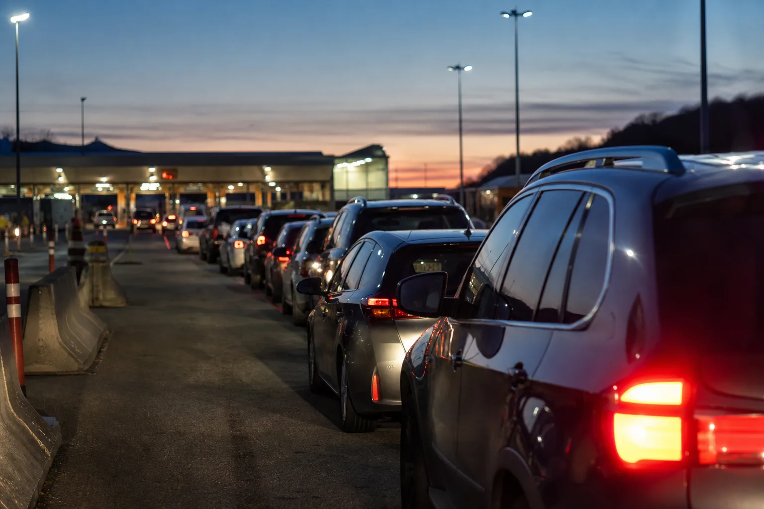 A queue of cars waiting at a land border crossing at dusk, tail lights glowing under floodlights.