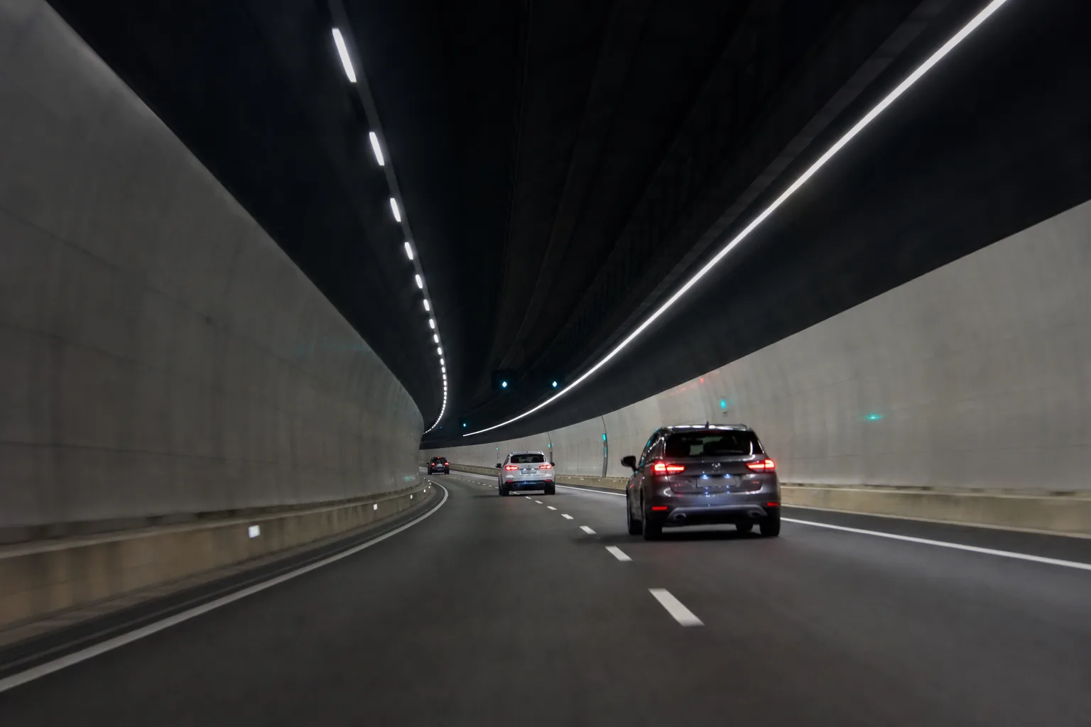 The lit interior of a road tunnel curving ahead, with ceiling light strips receding into the distance.