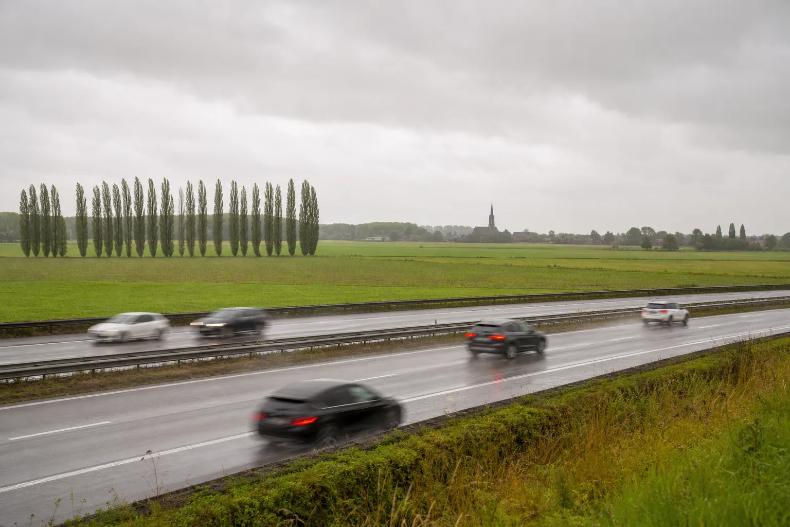 A wet Belgian motorway across flat Flemish farmland with a row of poplar trees and a distant church spire, evoking Belgium's speed limits.