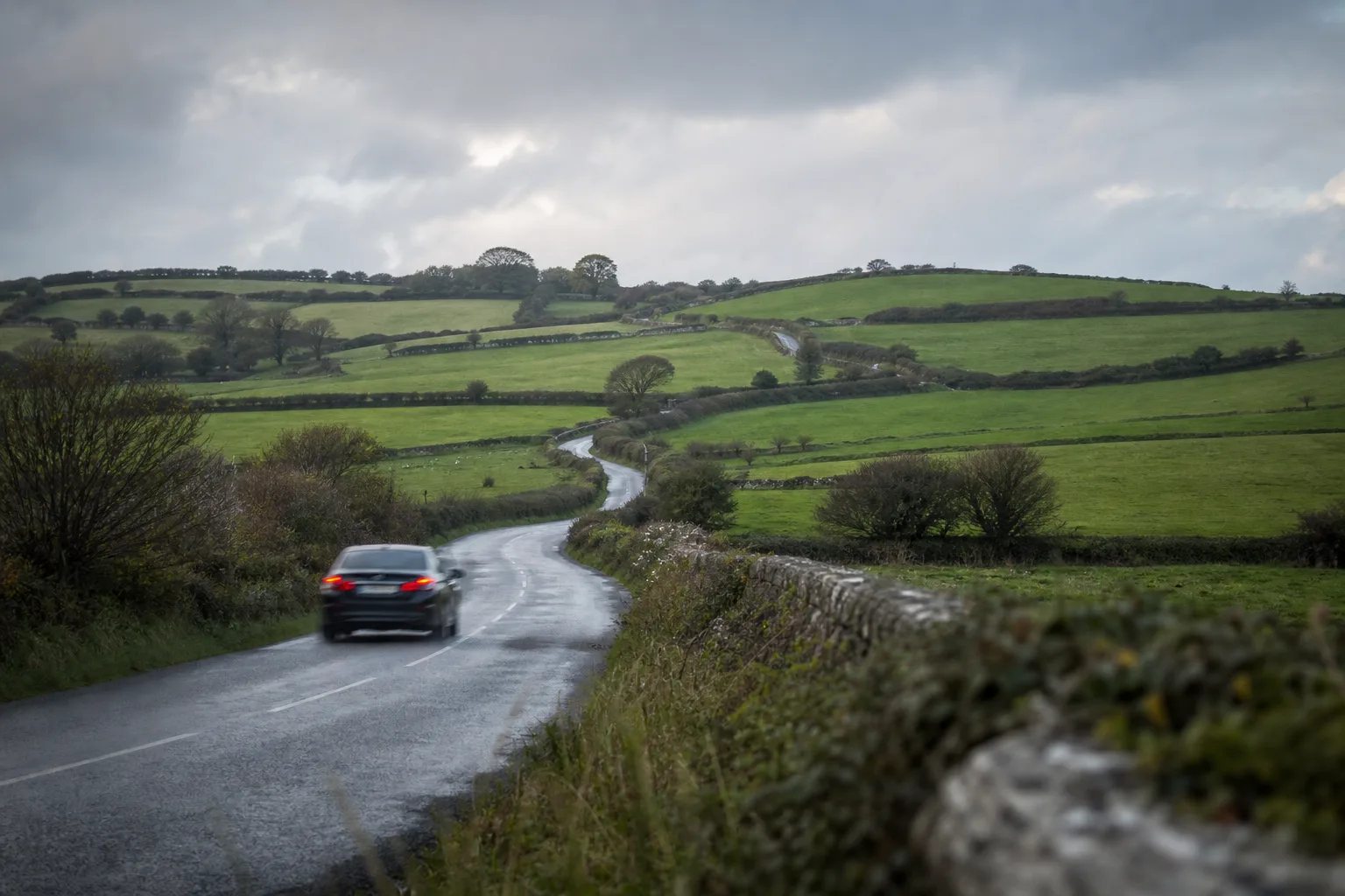 A car on a winding rural Irish road between hedgerows and stone walls across green hills under a grey sky, evoking Ireland's speed limits.