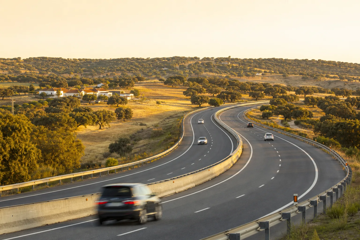 A Portuguese motorway curving through golden cork-oak countryside with white farmhouses at sunset, evoking Portugal's speed limits.