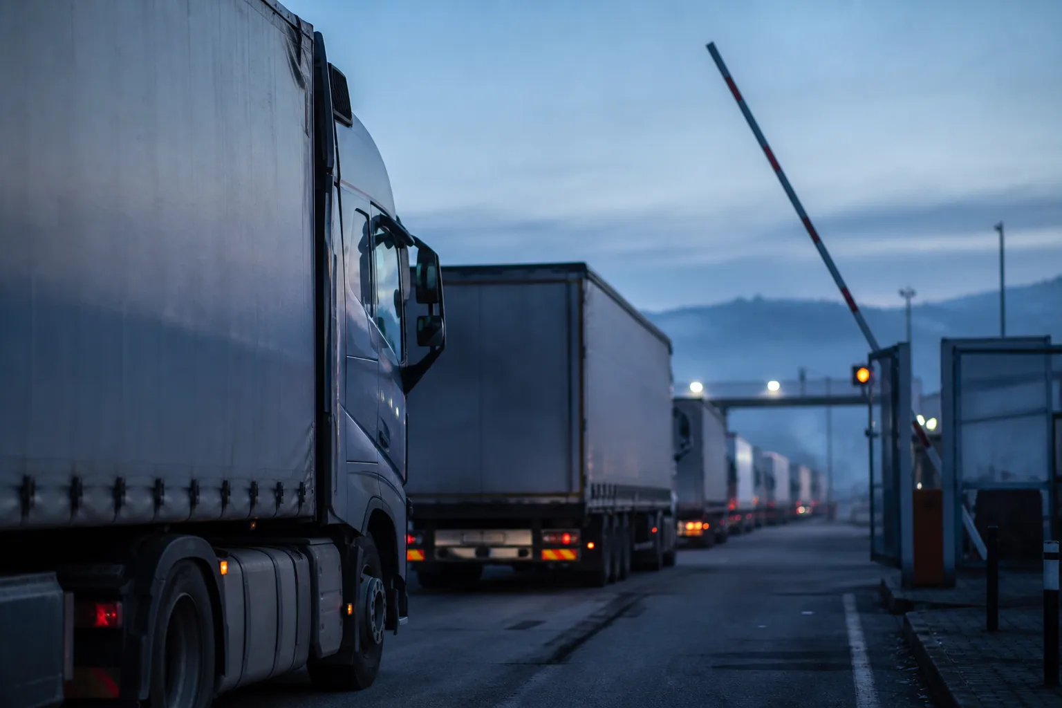 International articulated trucks queuing at a border crossing barrier at dawn.