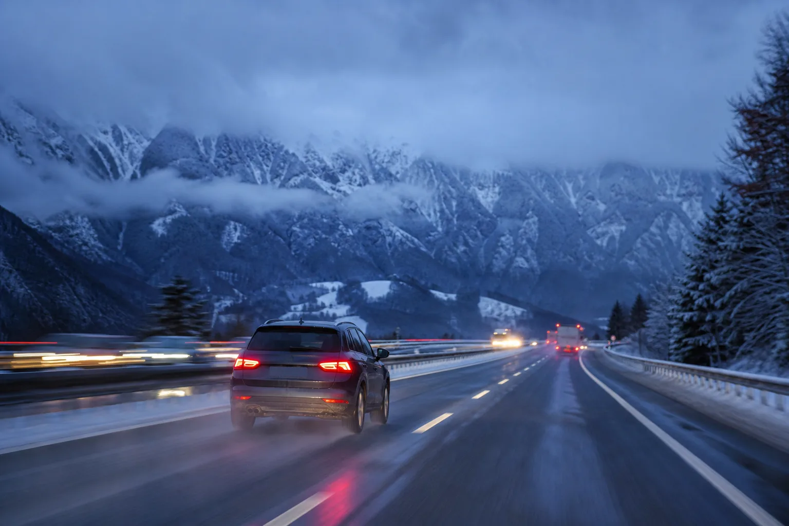 A car on a snow dusted alpine motorway at dusk, mountains behind and wet asphalt ahead.