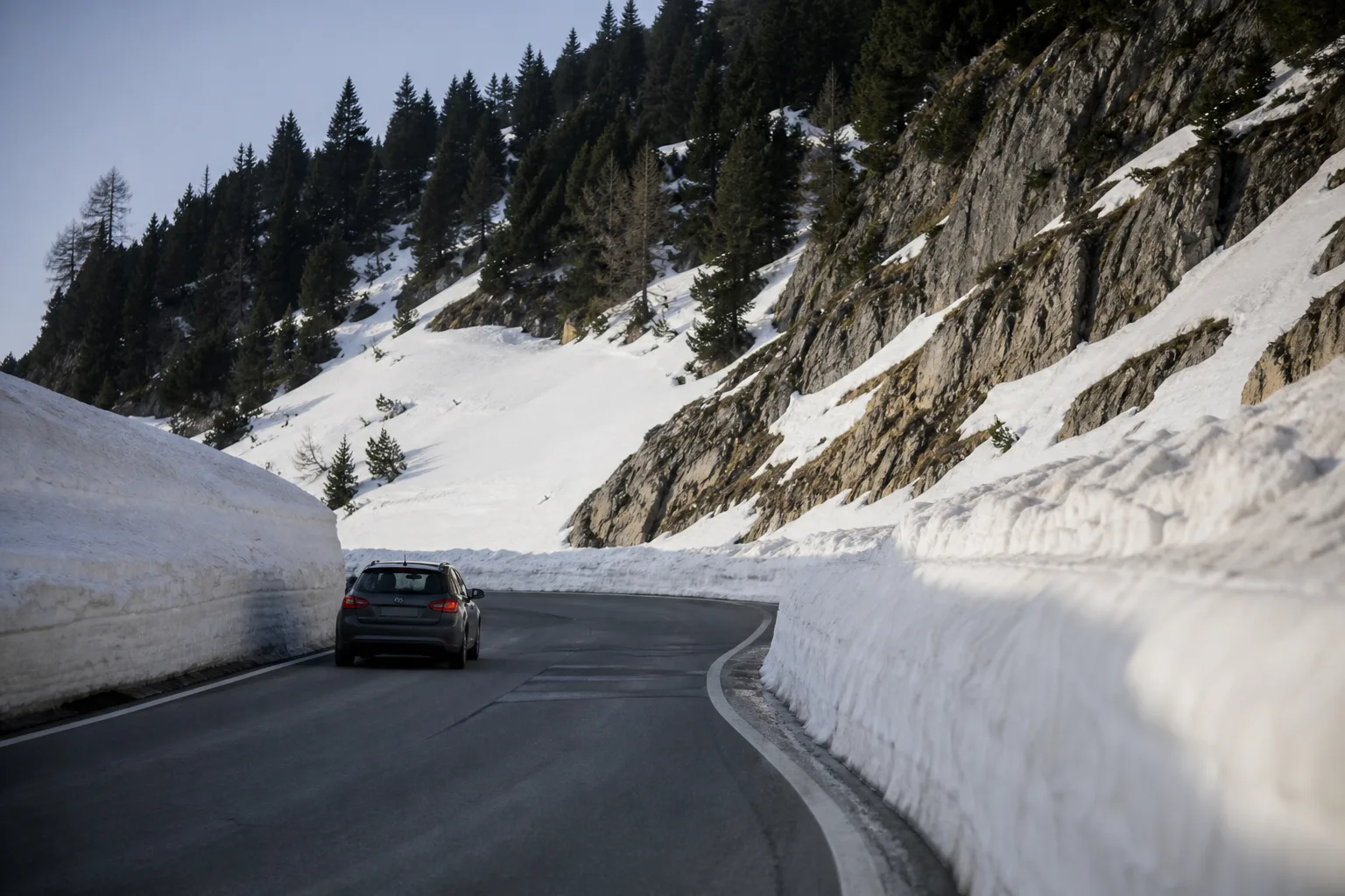 An Austrian alpine pass road between high snow banks with a car on a hairpin bend.