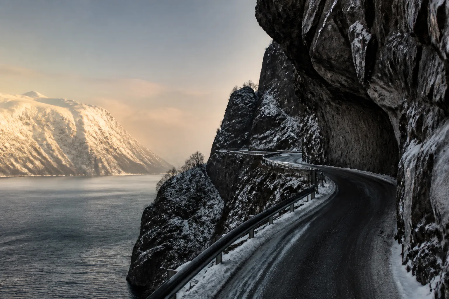A Norwegian fjord road cut into a snowy rock face above dark water.