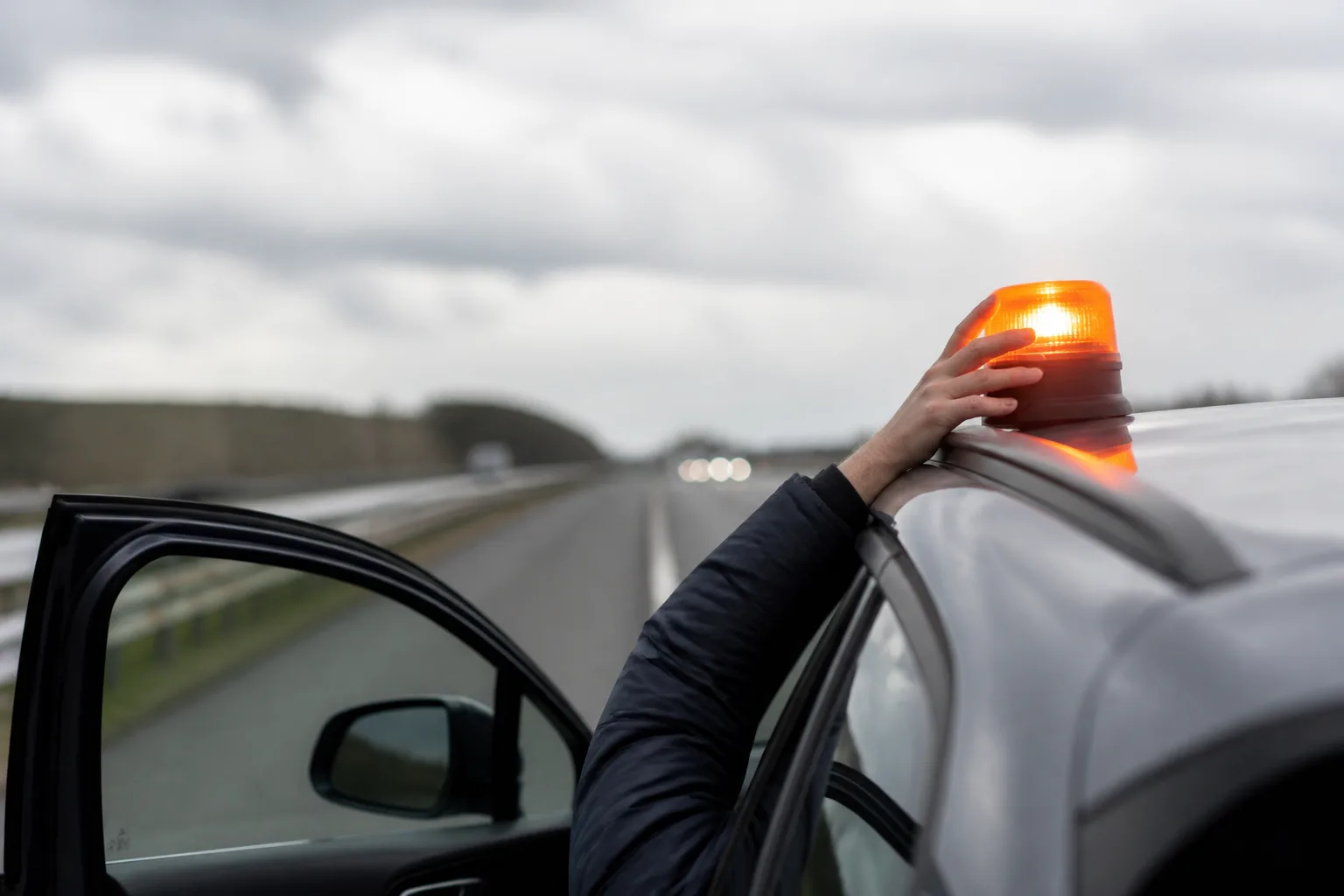 Eine Hand setzt aus der geöffneten Fahrertür heraus eine orangefarbene V16-Warnleuchte auf das Autodach, während der Wagen auf dem Standstreifen einer leeren Autobahn steht.