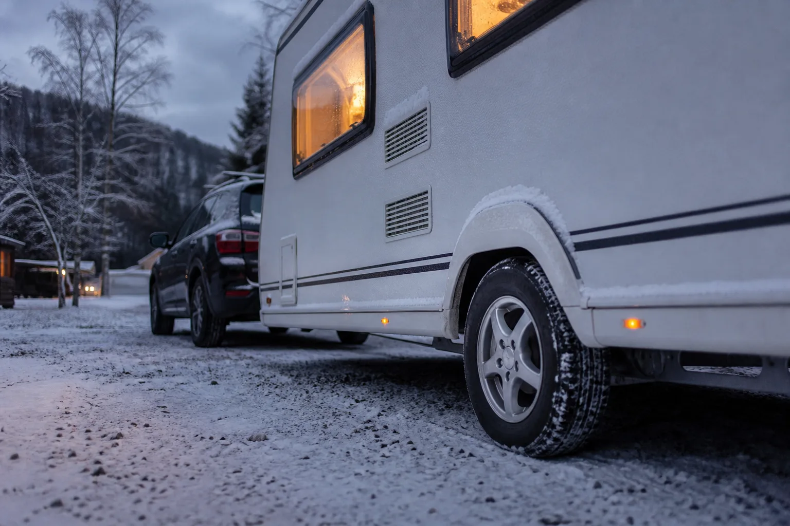 Wohnwagen mit verschneitem Reifen hinter einem SUV auf einem winterlichen Campingplatz in der Abenddämmerung.