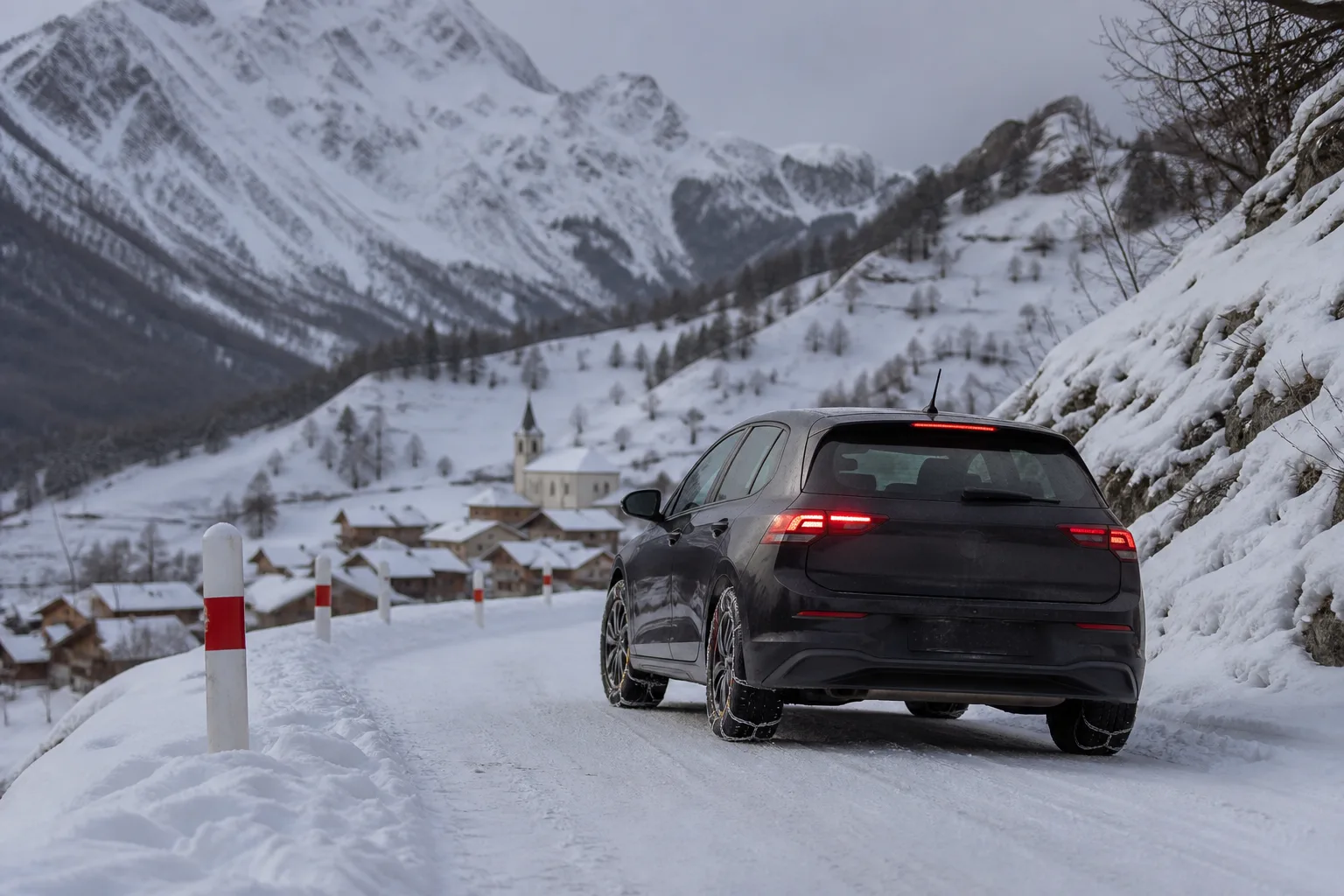 Auto mit Schneeketten an den Vorderrädern auf einer verschneiten Bergstraße in den französischen Alpen oberhalb eines Dorfes.