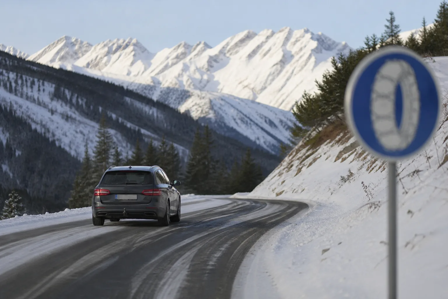 Kombi auf verschneiter Alpenpassstraße in Österreich, am Straßenrand ein blaues Schild zur Schneekettenpflicht.