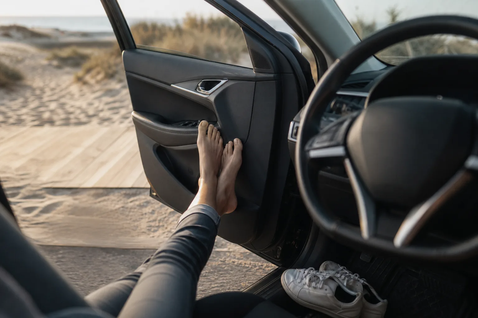 Bare feet resting outside an open car door at the beach while trainers wait on the driver's mat, the driving barefoot question in one scene.