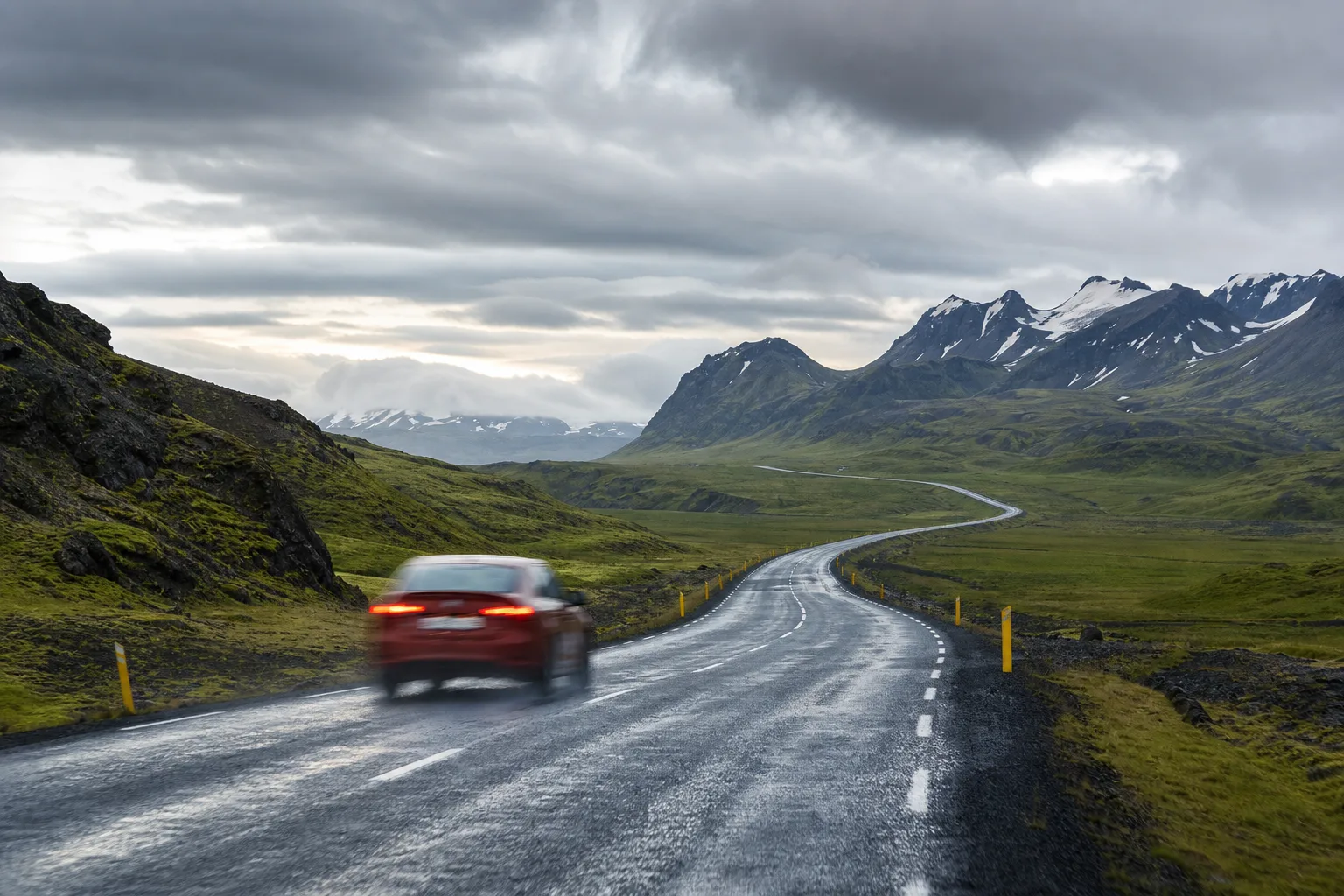 A car on Iceland's paved ring road winding through mossy hills toward snow-capped mountains under moody clouds, evoking Iceland's road speed limits.